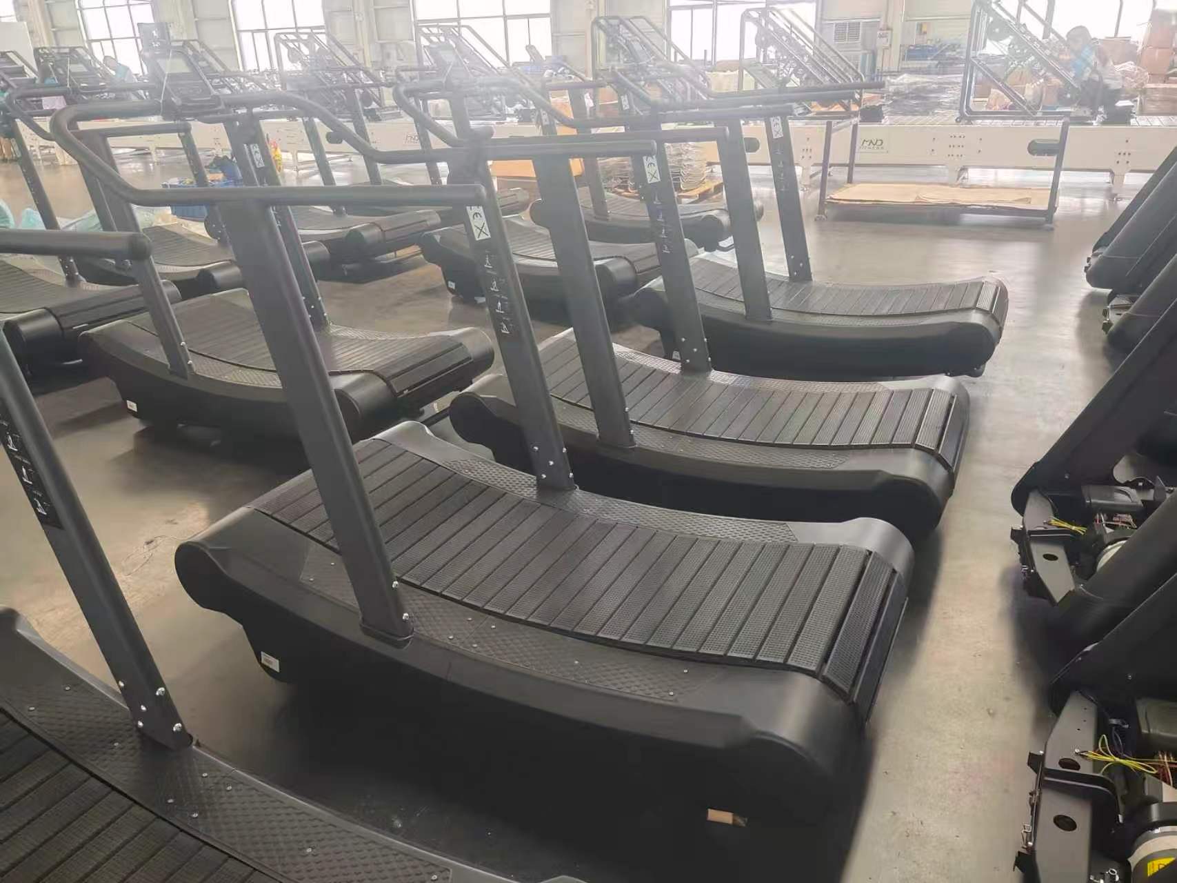 A row of black curved manual treadmills lined up on a production floor.